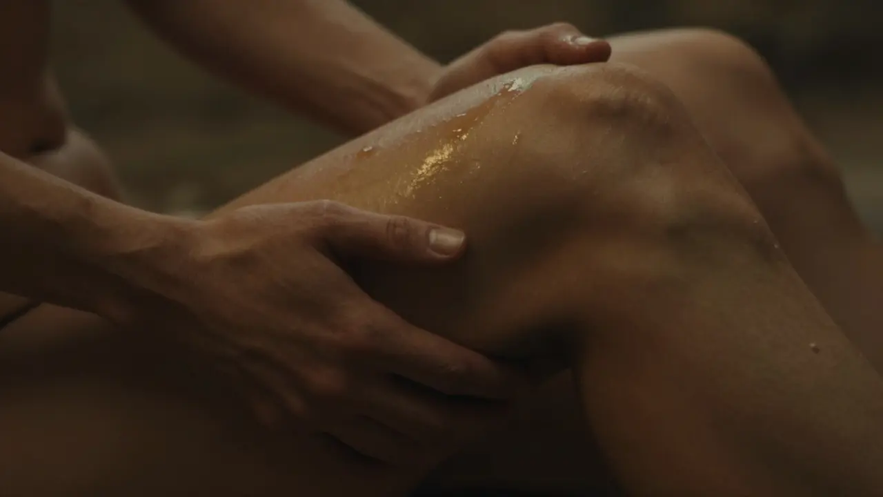 Two gentle hands touching a man’s inner thigh with care, oil shimmering under warm light, body blurred in soft focus.